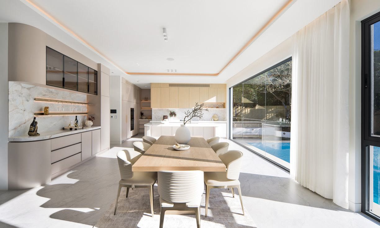 Open-plan dining area with a timber table and sand-coloured dining chairs beside a glazed corner window overlooking a pool