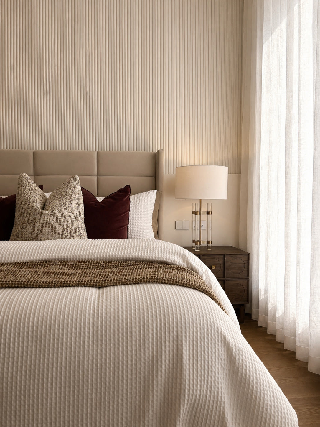 Bedroom with a quilted headboard, textured cream bedding, burgundy and linen cushions and a brass bedside lamp
