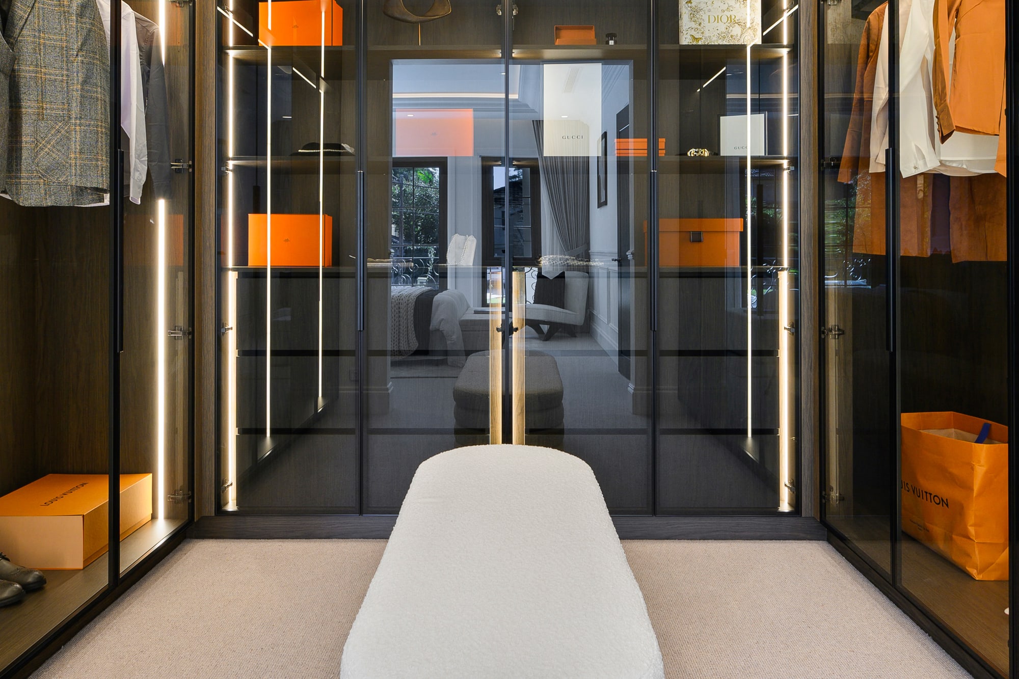 Upholstered ottoman beneath a crystal chandelier in a walk-in wardrobe with glass-fronted black joinery