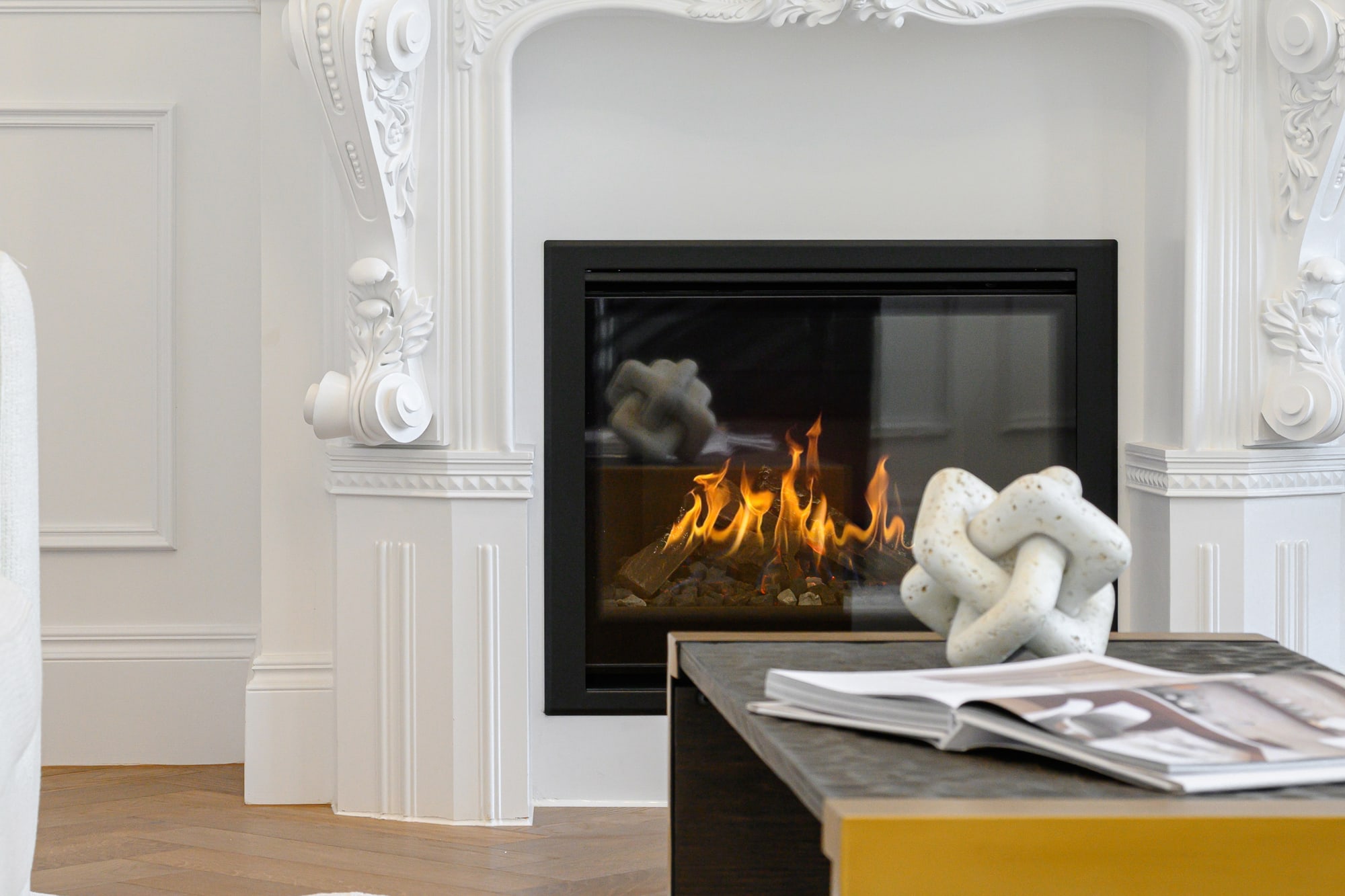 Ornate white fireplace mantel with a lit fire, sculptural ornaments and a black and mustard coffee table