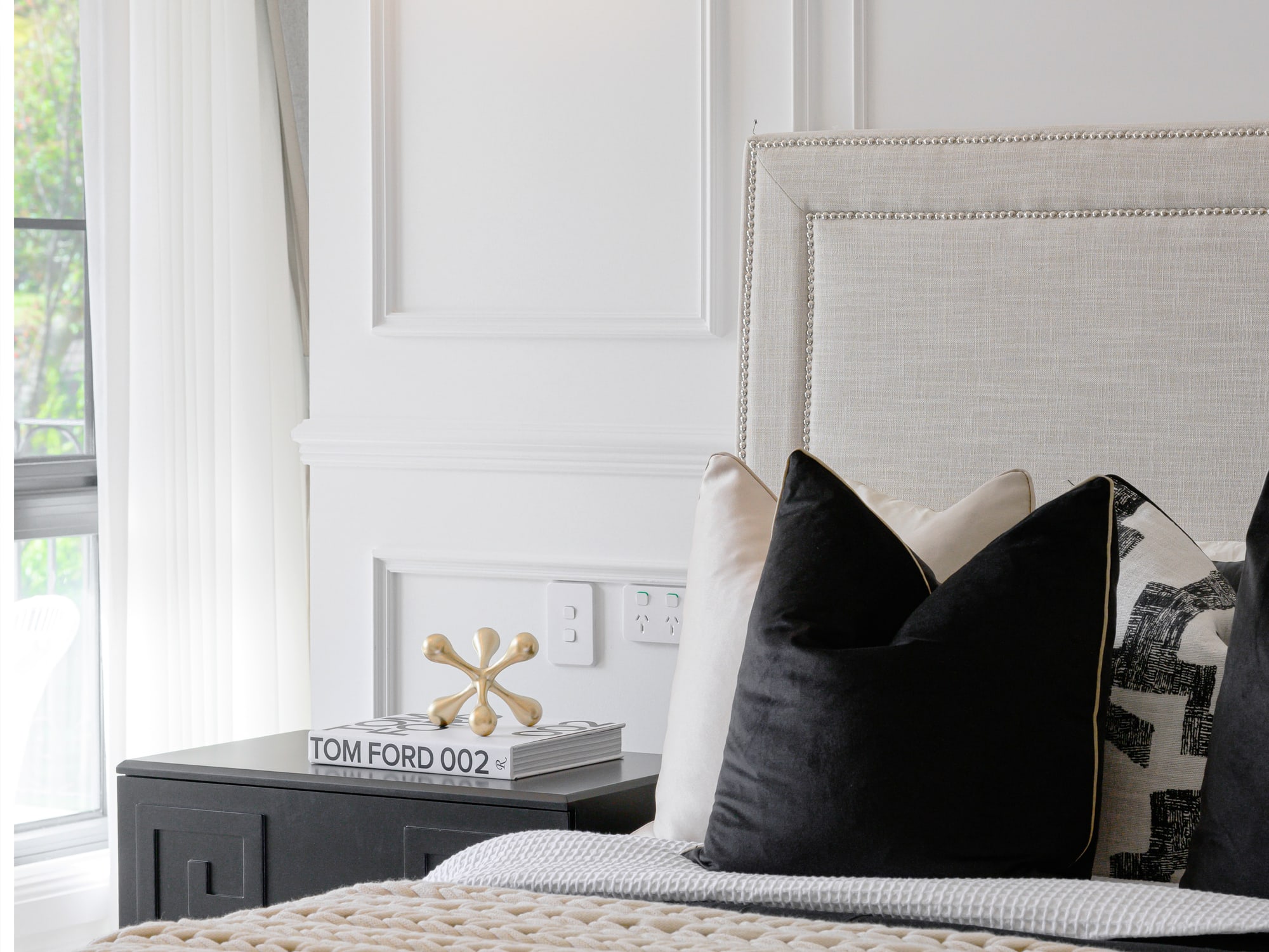 Bed dressed with black and cream cushions, a knitted throw and a pale upholstered bedhead