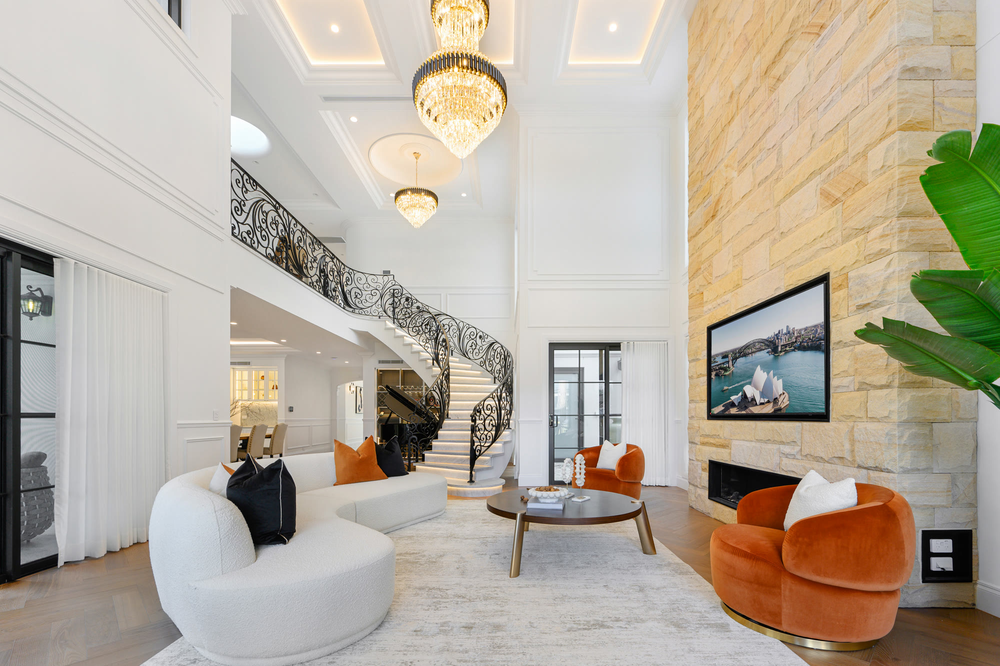 Double-height living room with curved cream sofas, orange velvet armchairs, a sandstone feature wall and a sweeping staircase with crystal chandeliers