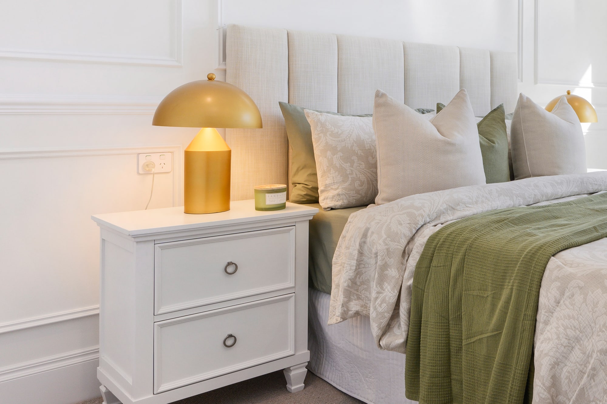 Gold dome lamp on a white bedside table beside a bed with a green throw and panelled walls