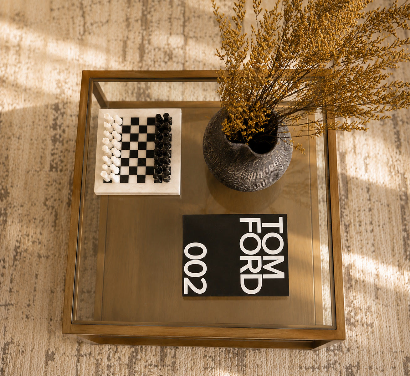 Glass coffee table styled with a chess set, dried flowers in a textured vase and a coffee-table book