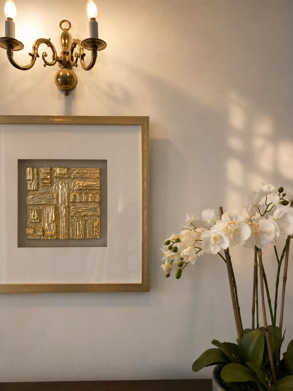 Gold textured artwork in a gilt frame beside a brass wall light and a white orchid
