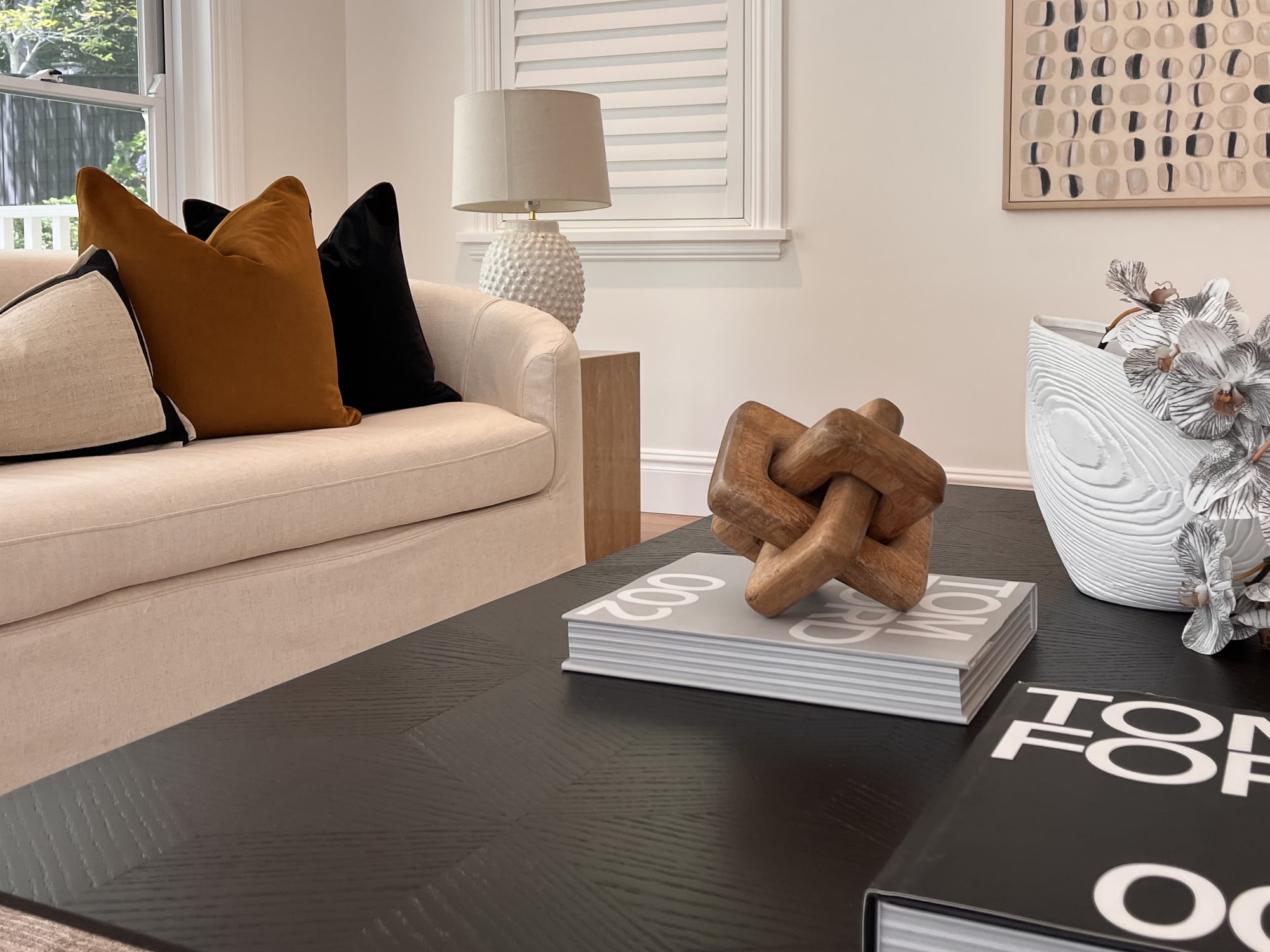 Black coffee table styled with books, a timber knot sculpture and a silver floral vase beside a cream sofa