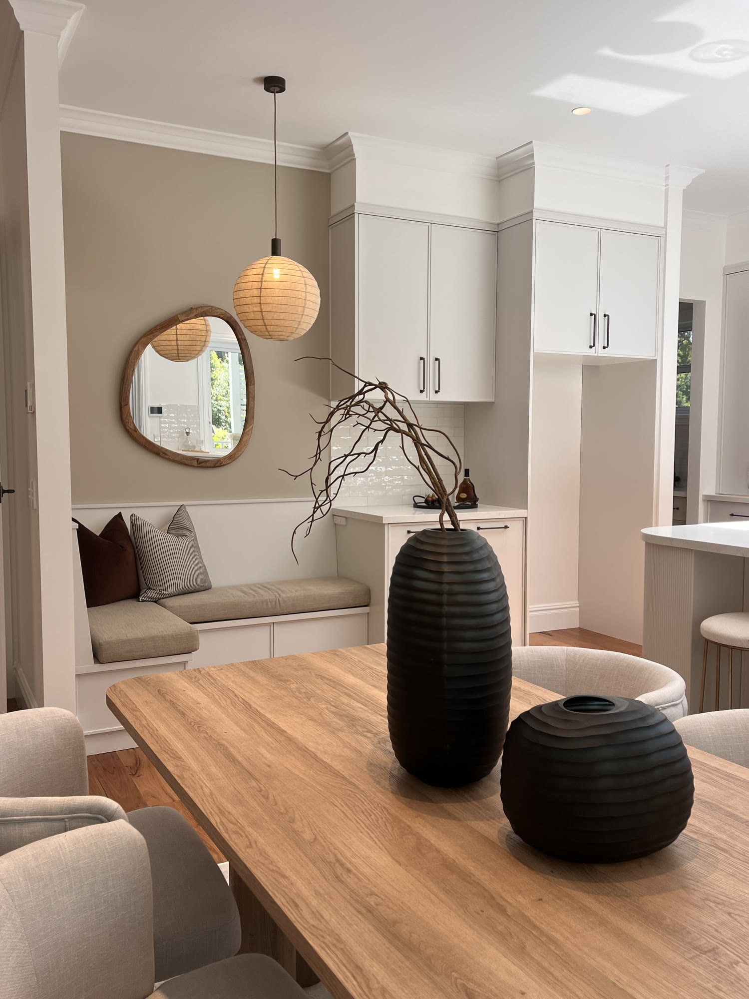 Timber dining table with black ribbed vases in front of a window seat, with a round mirror and kitchen beyond