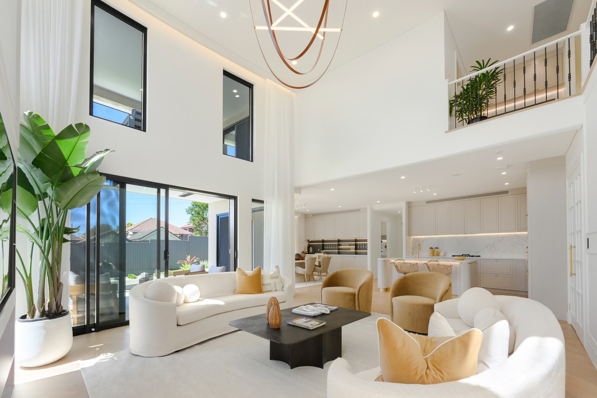 Open-plan living area under a double-height ceiling with curved cream sofas, a large indoor palm and the kitchen beyond
