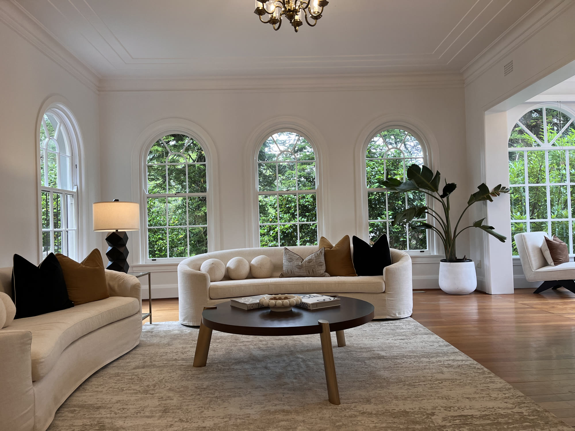 Wide living room with arched windows, curved cream sofas, a large round coffee table and a brass chandelier