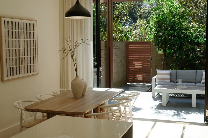 Dining table with a black pendant light and a vase of branches, with a courtyard seating area outside