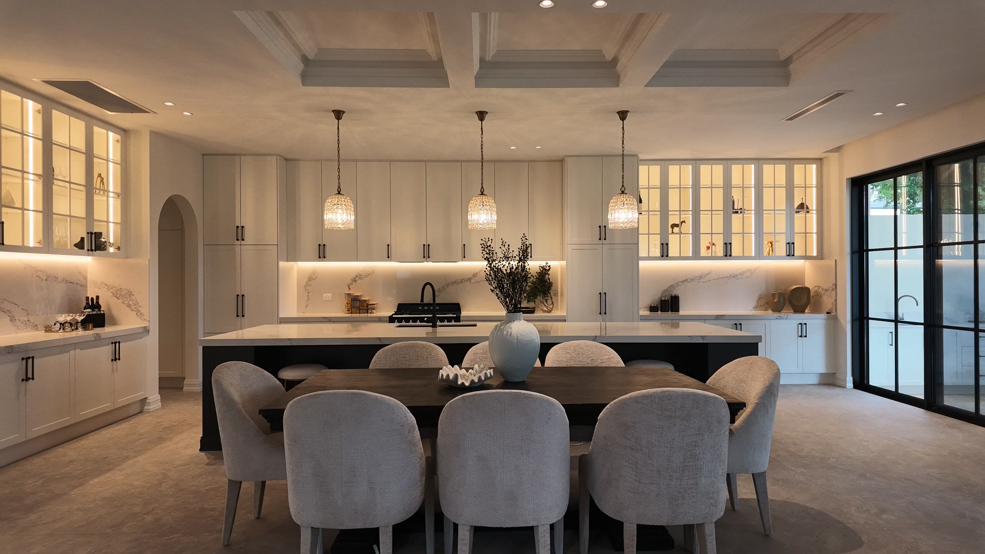 Open-plan dining and kitchen with a long black table, cream chairs and pendant lights under a coffered ceiling
