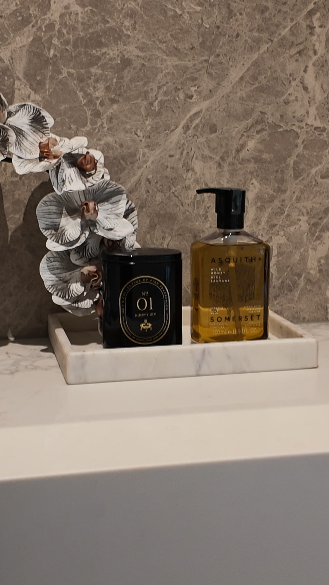 Candle, hand wash and a white orchid on a marble tray against a stone splashback